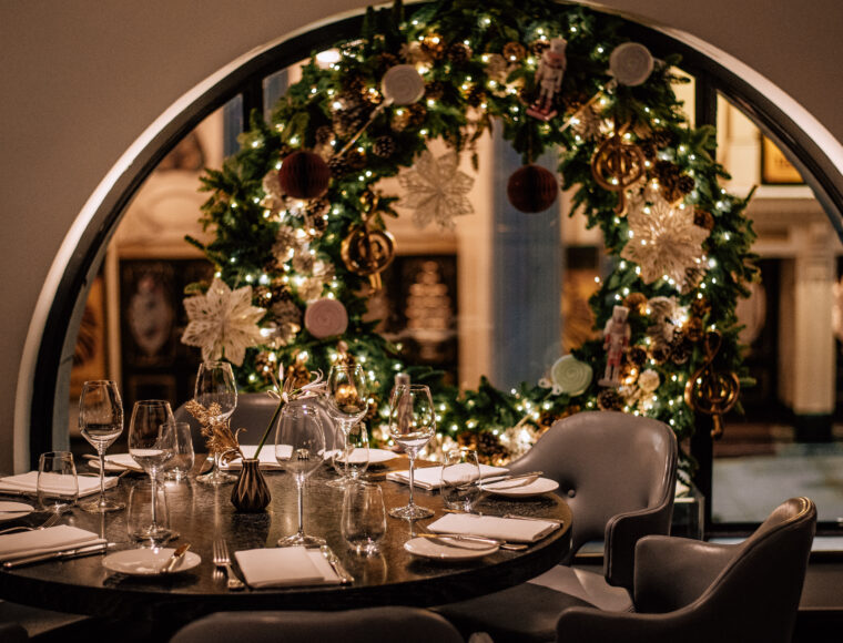 Restaurant table set with water and wine glasses, knives, forks, and napkins; in the background, a window decorated with a lit Christmas wreath glows warmly against the nighttime sky.