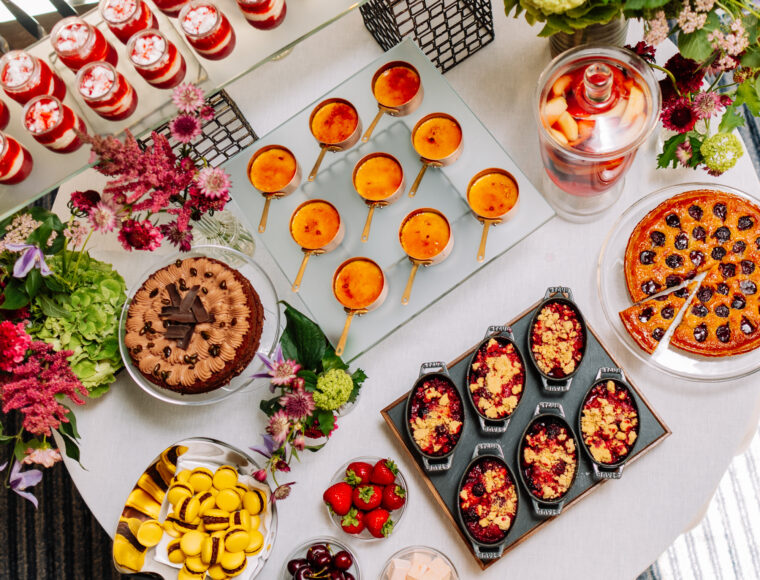 Aerial view of a decorated dessert buffet table adorned with flowers, displaying a variety of desserts including macarons, crème brûlée dishes, parfaits, fresh fruits, cakes, pastries, and poached plums.