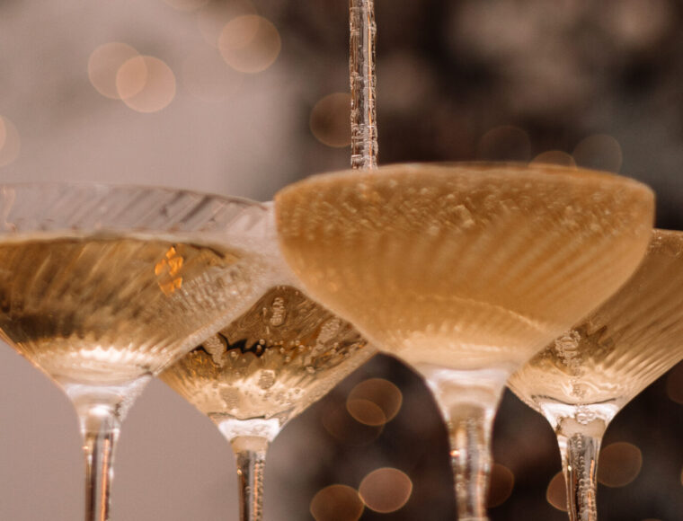 A champagne tower with sparkling Christmas lights softly blurred in the background; a bottle of champagne is being poured into the top glass, causing it to cascade down the tower.