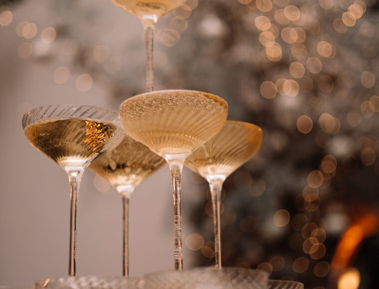 A champagne tower with sparkling Christmas lights softly blurred in the background; a bottle of champagne is being poured into the top glass, causing it to cascade down the tower.