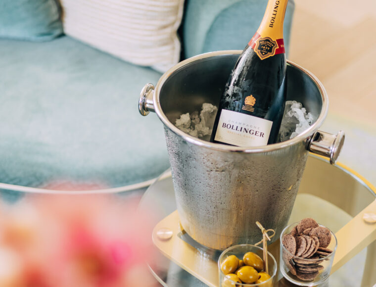 Bottle of Bollinger champagne chilling in an ice bucket on a table next to a blue sofa in a hotel room; olives and crackers are arranged on the table beside the champagne. Soft-focus pink flowers blurred in the corner of the image.
