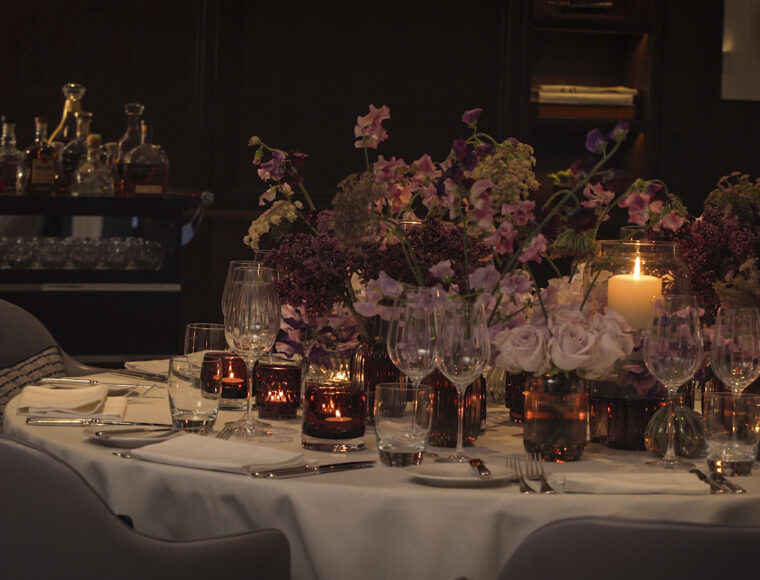 Elegant dining table set with white tablecloth, silverware, wine glasses, candles, and a large floral centerpiece of purple and pink flowers, in a dimly lit, high-end restaurant setting.