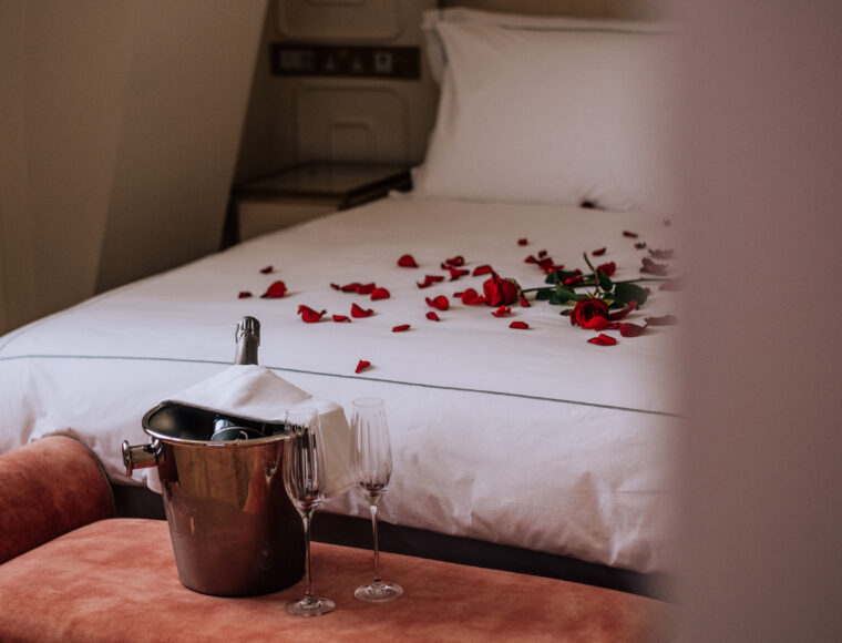 A neatly made bed with white linens, scattered red rose petals, and a champagne bottle chilling in a silver ice bucket beside the bed, creating a romantic atmosphere.