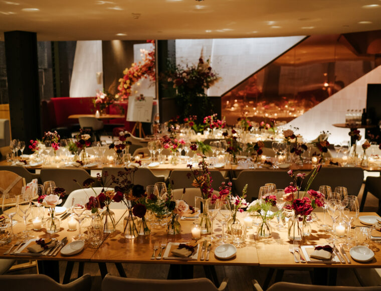 Long wooden tables set for a formal dinner, adorned with floral centerpieces of red and pink blooms in small vases, candles, glassware, and place settings; warm lighting creates an elegant atmosphere.