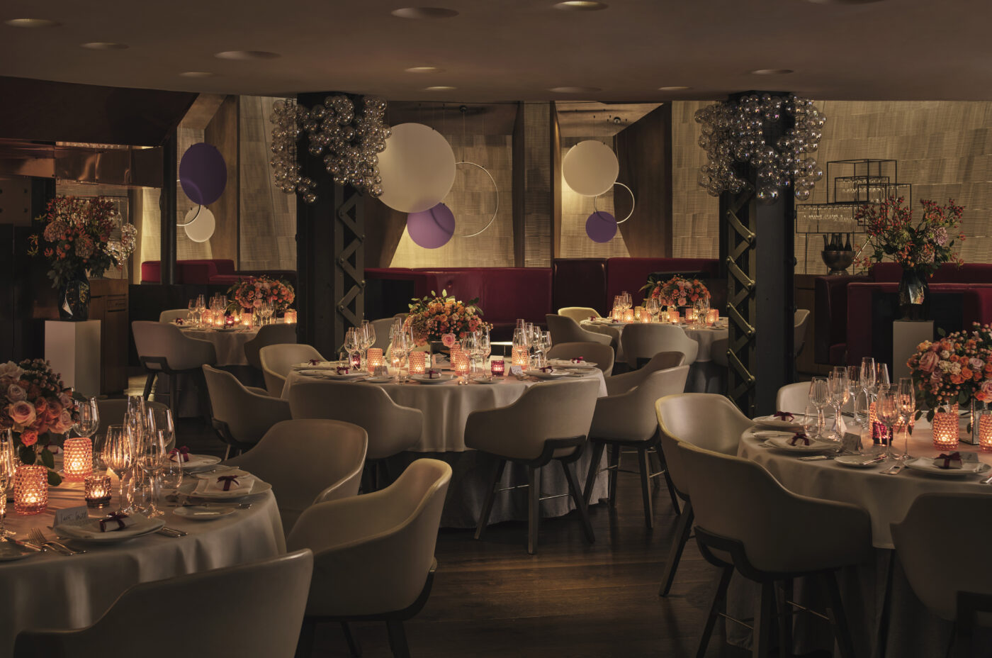Elegant restaurant interior at night, multiple round tables set for a formal event, featuring white linens, glassware, candles, and floral centerpieces; soft lighting illuminates the room with decorative globe chandeliers and circular hanging art.