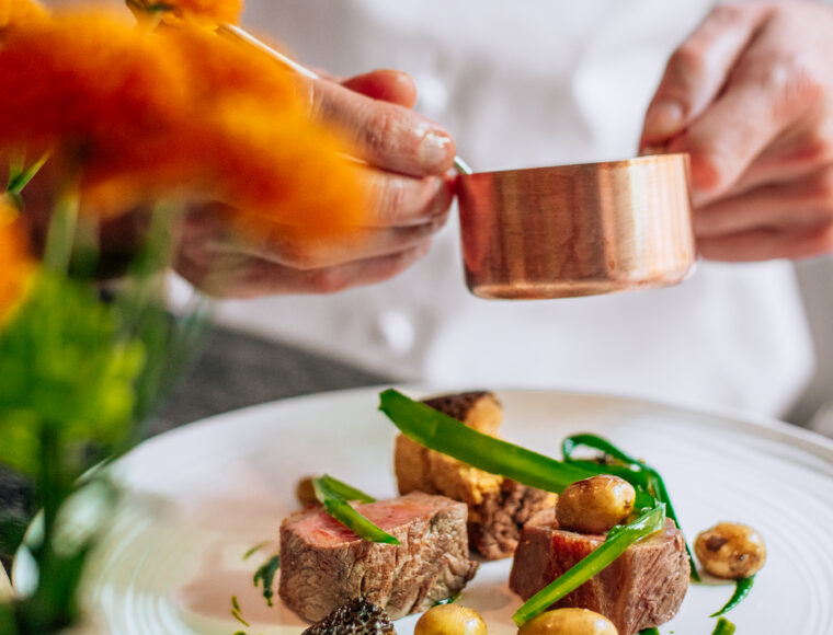 A restaurant chef pours sauce from a copper pot onto a luxury plated dish featuring meat, potatoes, morel mushrooms, and green garnish, with vibrant orange flowers blurred in the foreground.