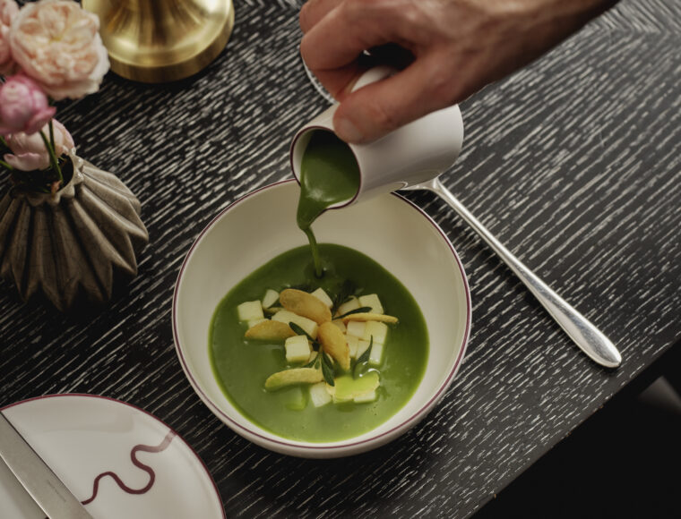 Chef pours vibrant green herb soup into a bowl topped with diced vegetables and crisp garnishes on an elegant restaurant table at One Aldwych’s Indigo restaurant in London, Covent Garden.