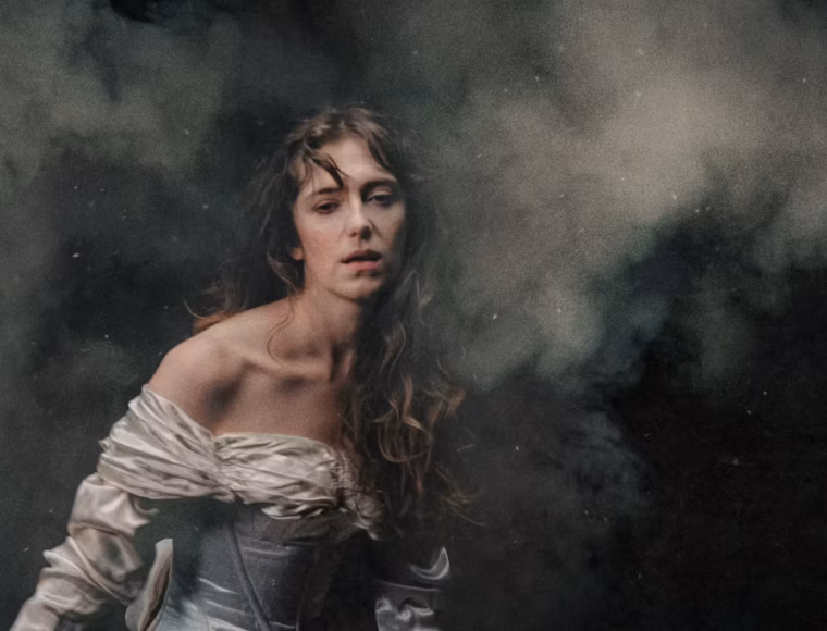 Royal Opera House perfomrance poster in Covent Garden: A person with long, flowing hair wears a white off-the-shoulder dress while partially submerged in dark, murky water, creating a moody and atmospheric ethereal portrait.